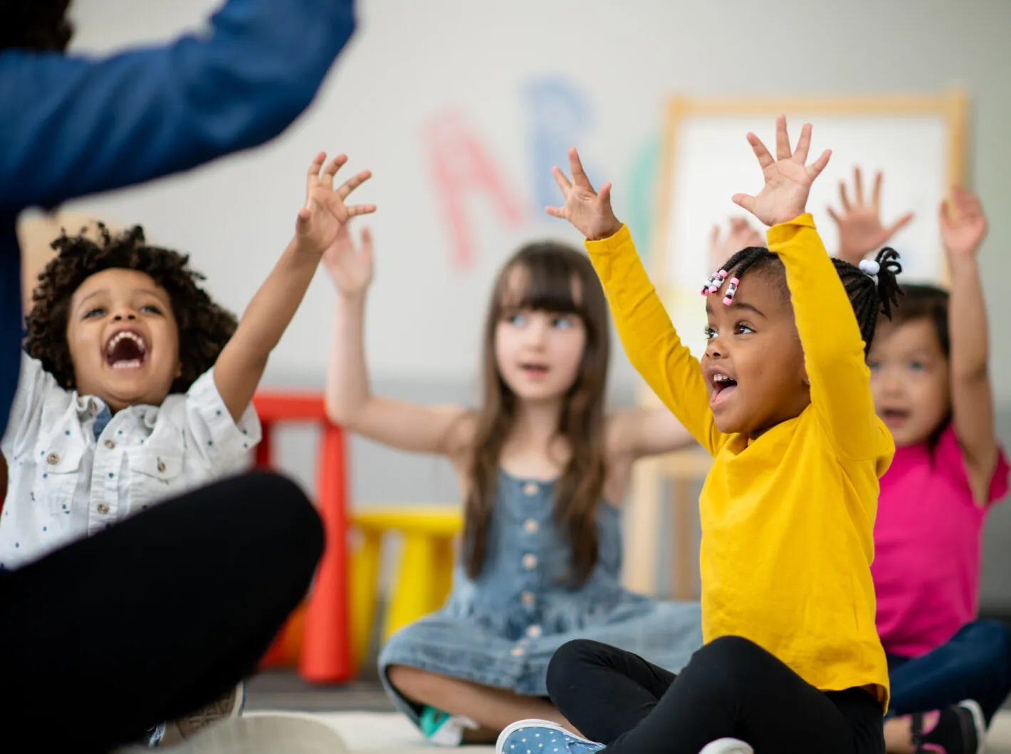 Children sitting on the floor, joyfully raising hands in a classroom.