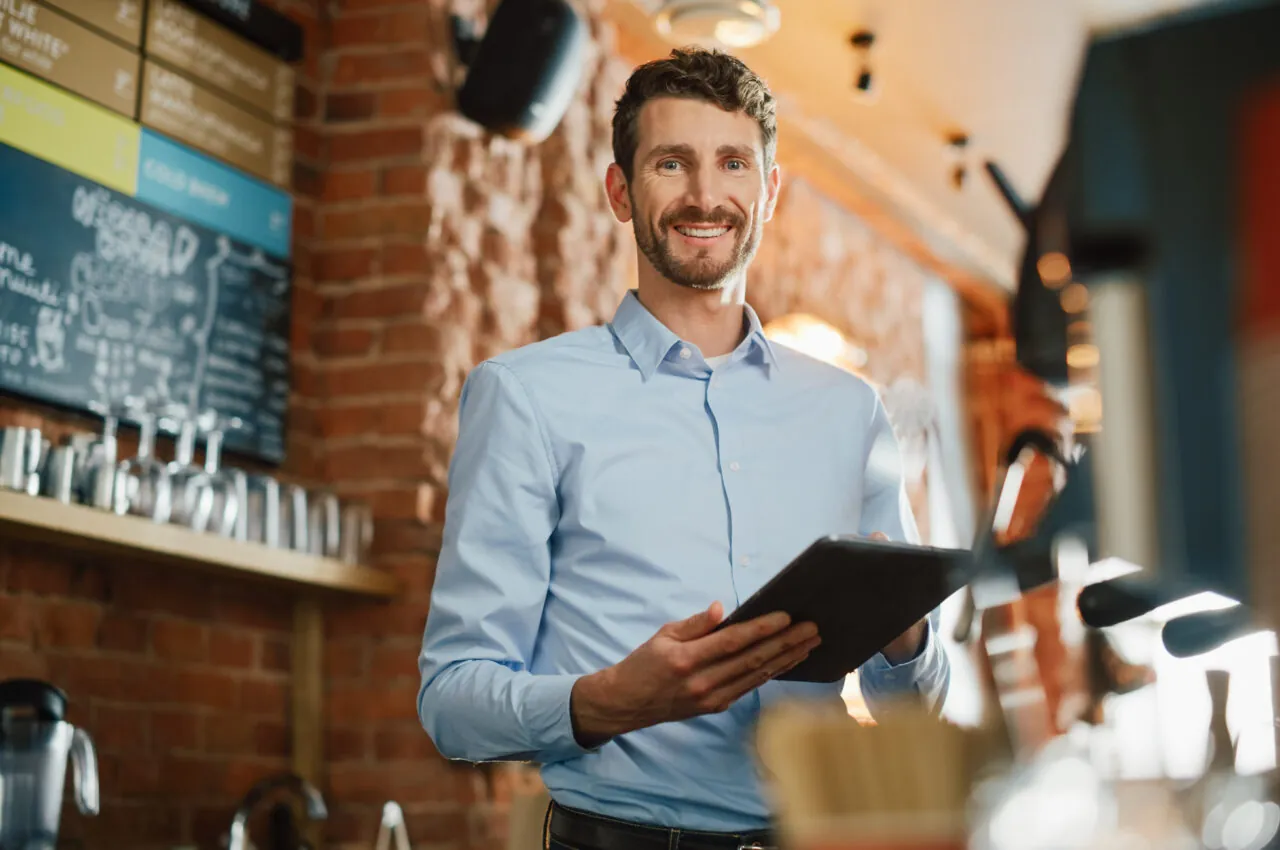 GettyImages-1293175092.jpg: Smiling man in a blue shirt holding a tablet in a cafe with a menu board behind him