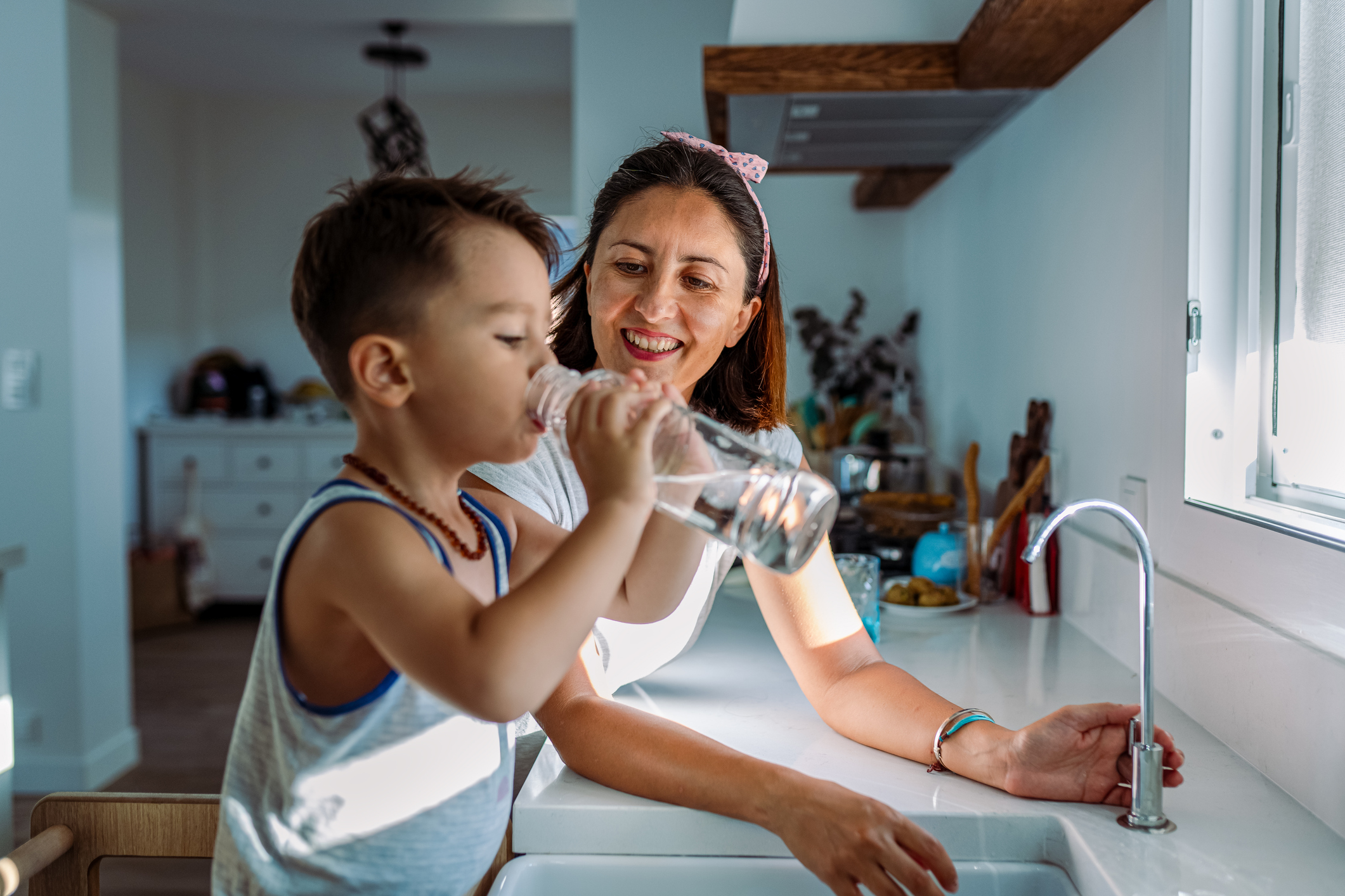 Child drinking water from a bottle, smiling adult nearby in kitchen.