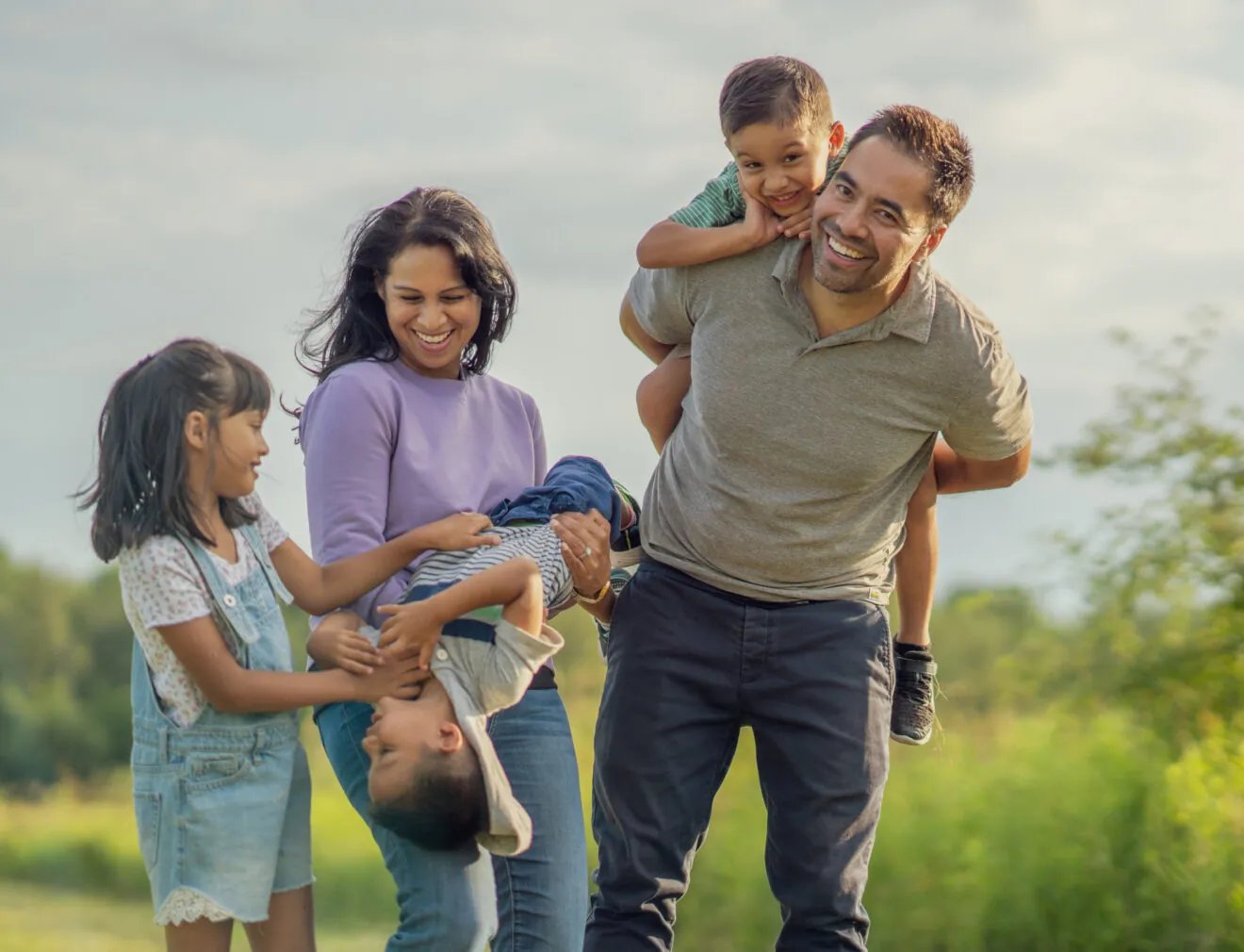 Family playing joyfully outdoors, parents carrying children playfully, sunny day, green field in background