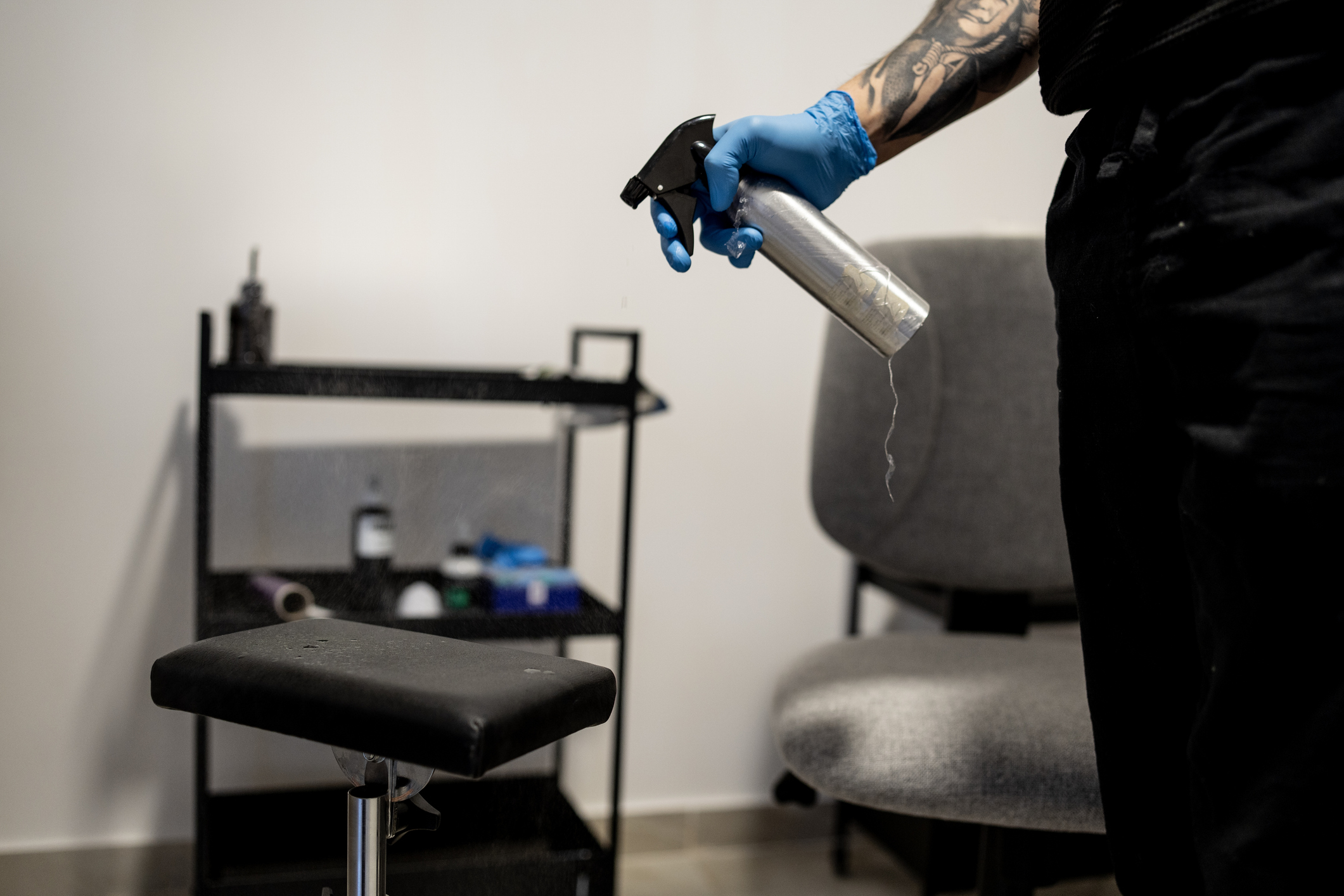 Tattoo artist cleaning stool with spray bottle, wearing gloves.