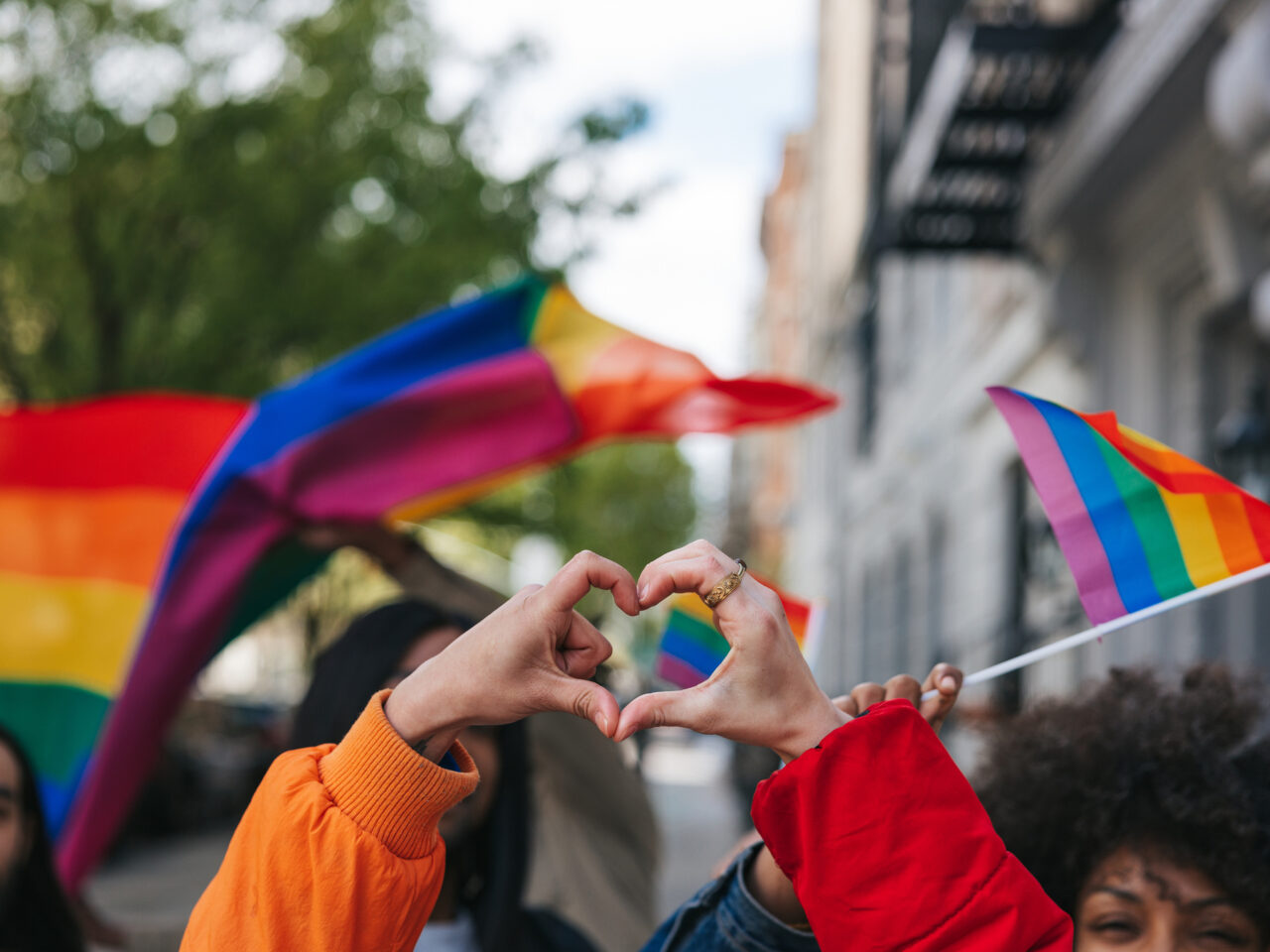 Hands forming a heart shape, rainbow flags waving in the background.