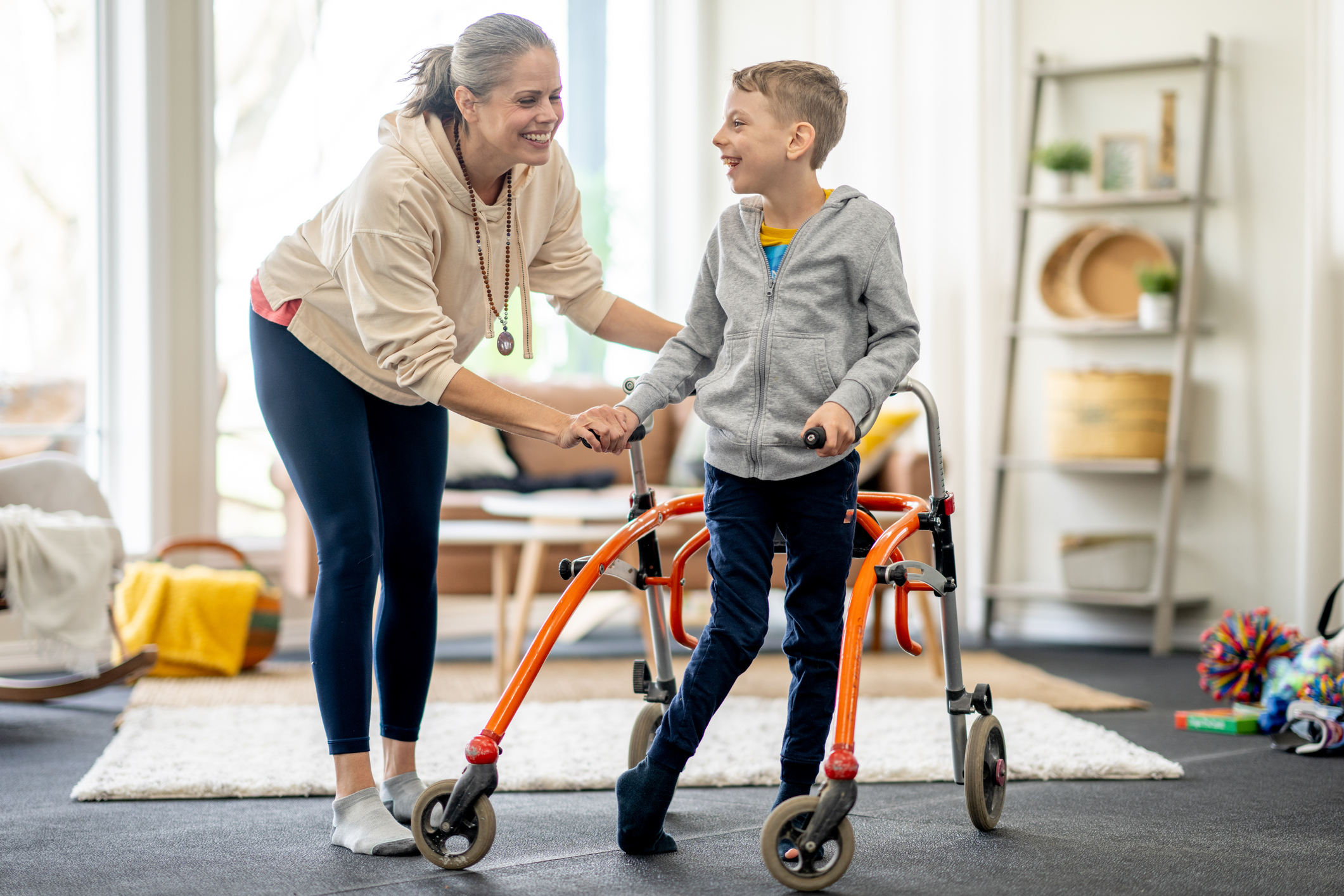 Photo of an adult and child smiling, child using an orange walker in a cozy room.