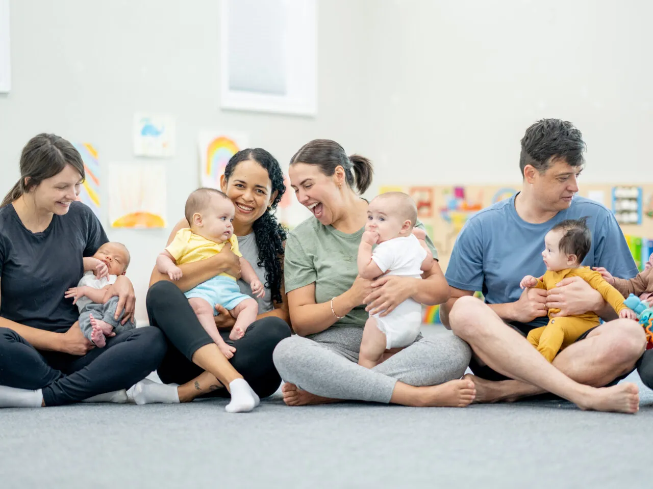 Adults sitting in circle with babies, happily interacting in a bright room with children's artwork on walls.