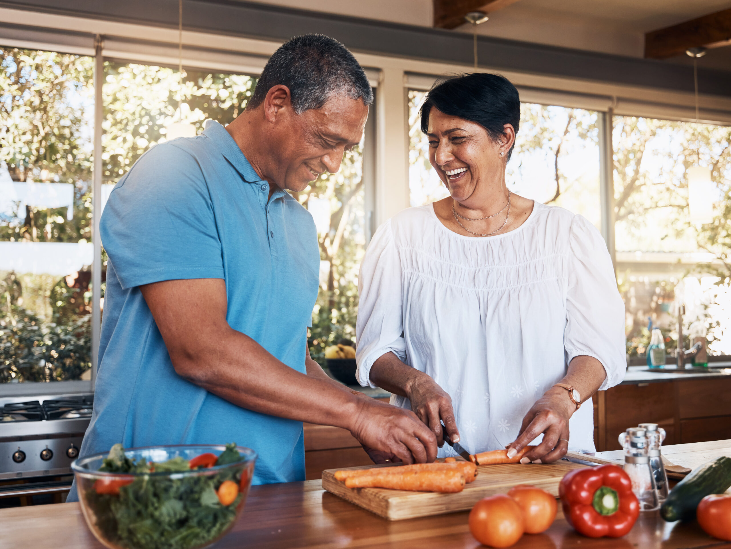 Couple laughing together while cutting carrots on a wooden board in a sunlit kitchen