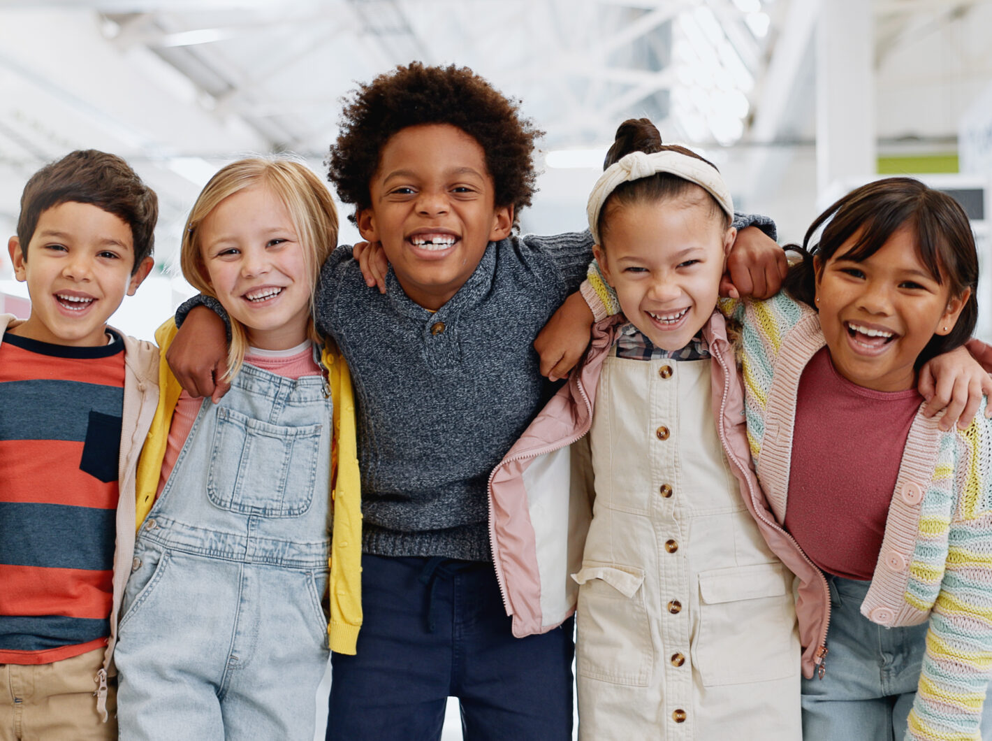 Five children smiling, standing arm in arm indoors, expressing joy and friendship.