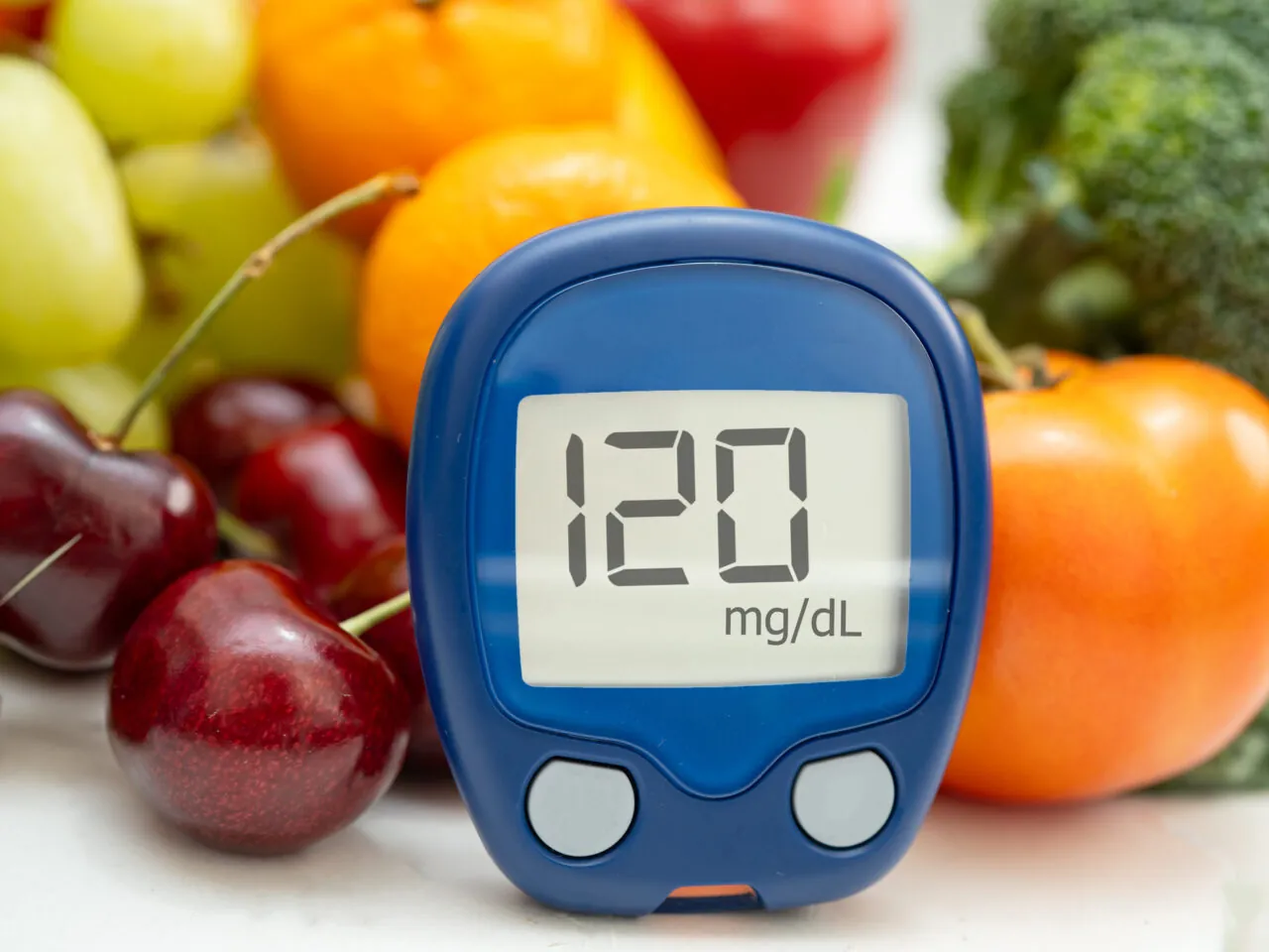 Glucometer displaying 120 mg/dL surrounded by fruits and vegetables.