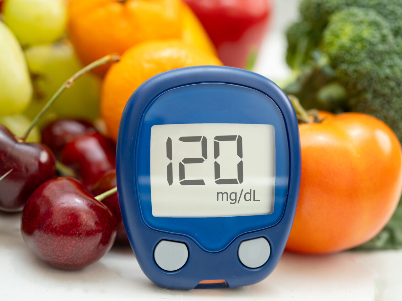 Glucometer displaying 120 mg/dL surrounded by fruits and vegetables.