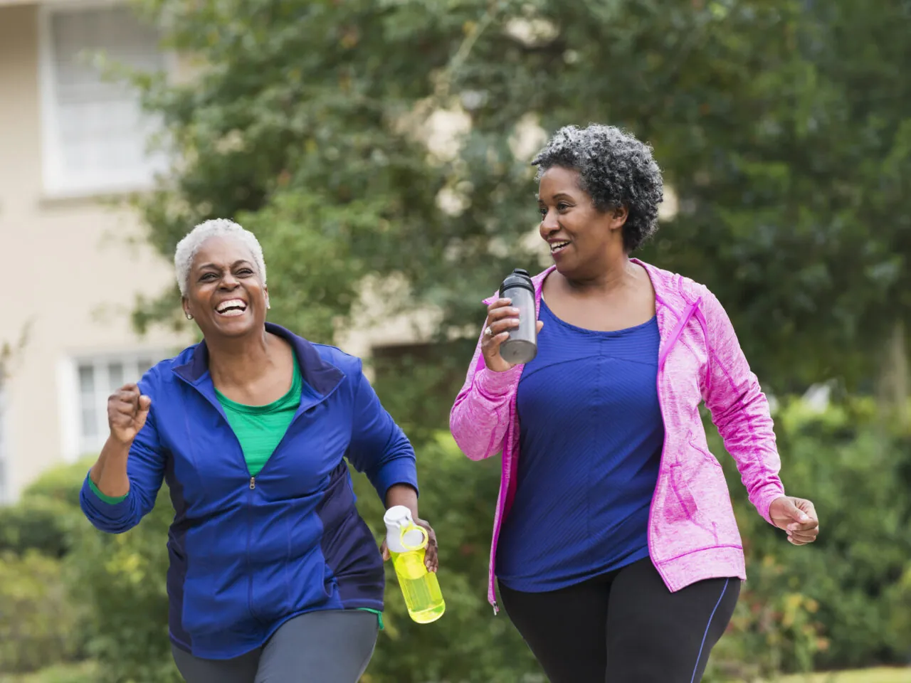 Two women wearing athletic clothing enjoy a walk outdoors, smiling and holding water bottles.