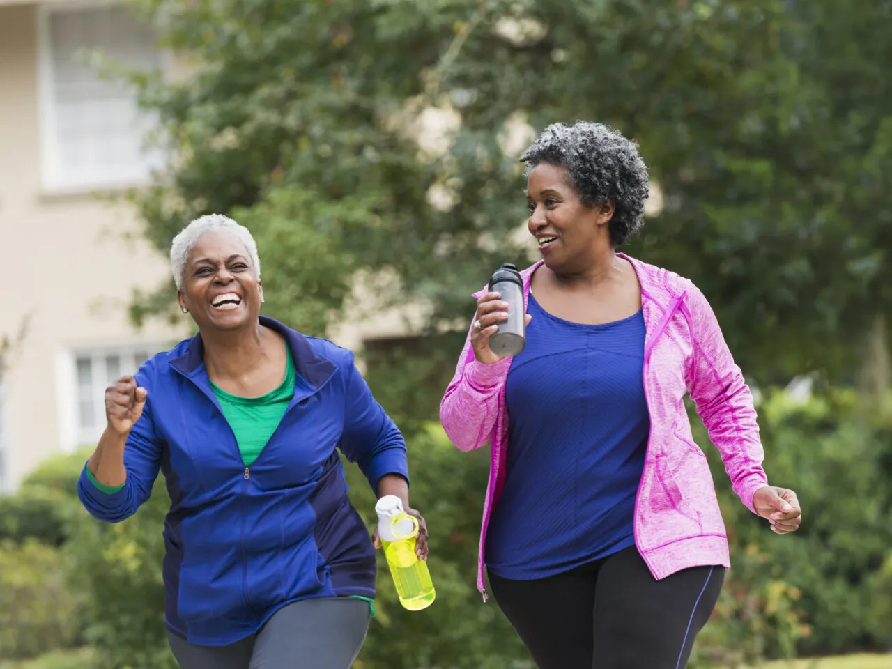 Two women wearing athletic clothing enjoy a walk outdoors, smiling and holding water bottles.