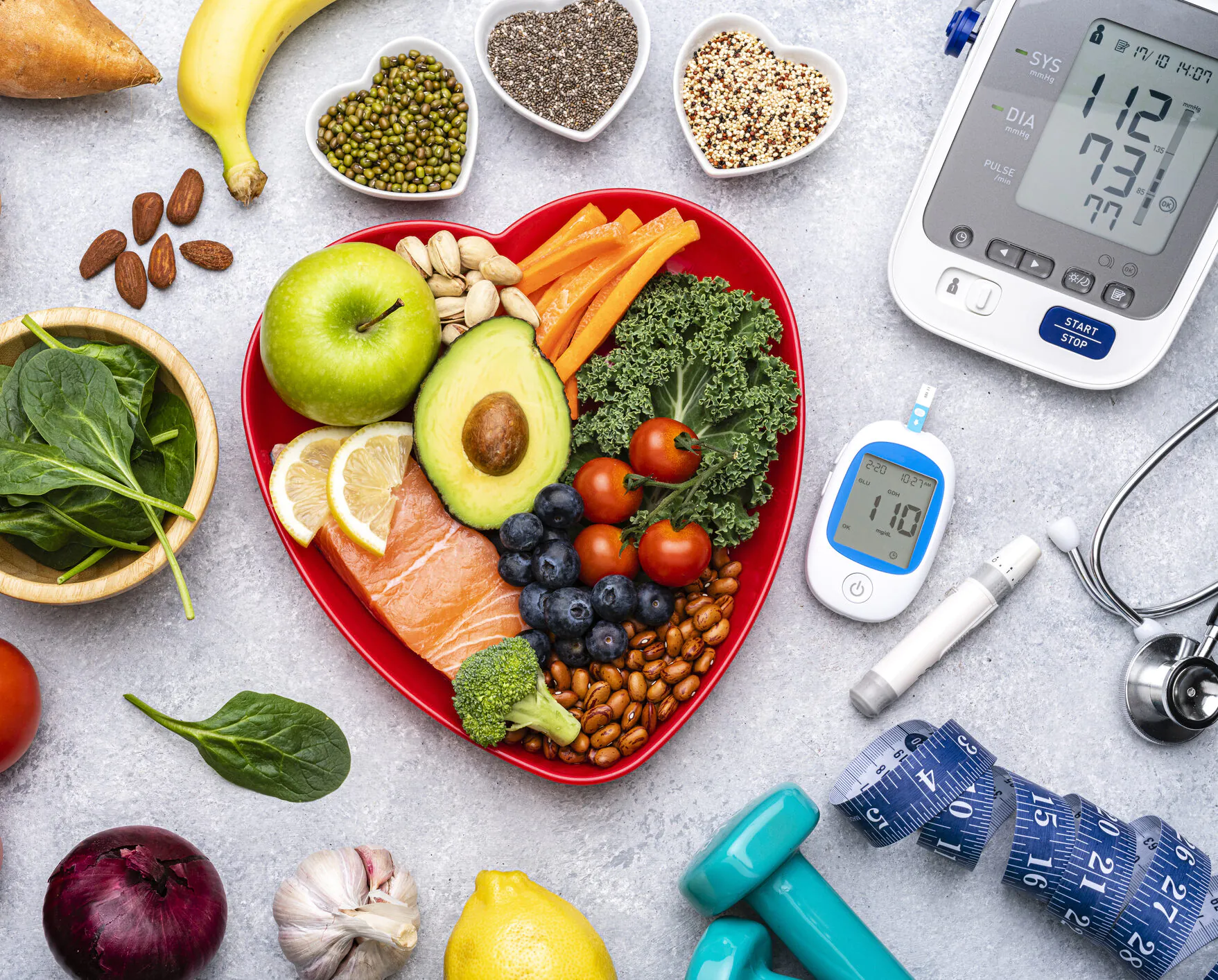 Heart-shaped bowl with vegetables, fruits, nuts, and salmon beside a blood pressure monitor and glucose meter