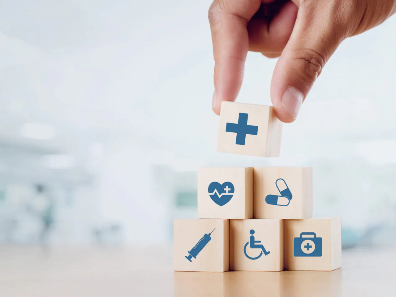Hand stacking healthcare-themed blocks: cross, heart, pills, syringe, wheelchair, first aid.