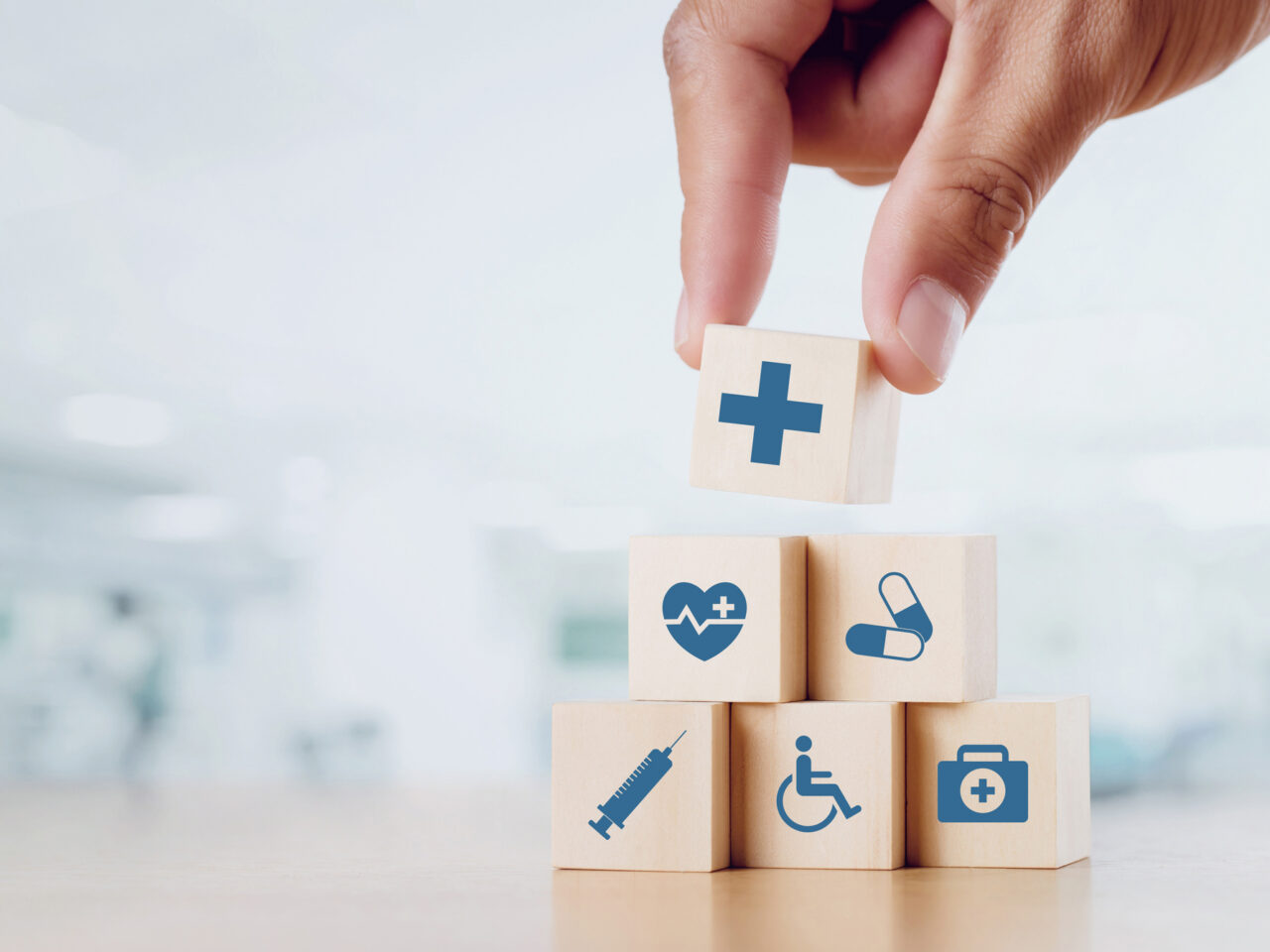 Hand stacking healthcare-themed blocks: cross, heart, pills, syringe, wheelchair, first aid.